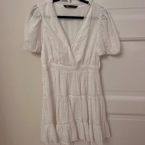 Vit kort klänning från Zara - Vit kort klänning från Zara med v-ringning, puffärmar och broderade detaljer. Använd 1 gång på midsommar. Säljer för det blivit för kort. Storlek M men är mer åt S-hållet 💗