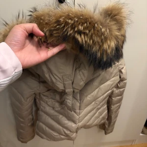 Beige pufferjacka HOLLIES med päls - Säljer en beige pufferjacka med fluffig päls på huvan. Jackan har quiltad design och är perfekt för kalla dagar. Färgen är neutral och passar till det mesta. Huvan har snygg pälsdetalj i bruna och svarta toner. Jackan har långa ärmar och dragkedja framtill.