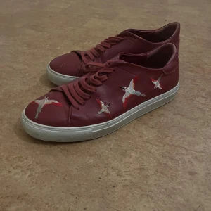Röda sneakers från Axel Arigato - Snygga röda sneakers från Axel Arigato i skinn med broderade fåglar på sidorna. Skorna har vita sulor och matchande röda skosnören. Perfekt för dig som vill sticka ut med en unik detalj på fötterna. Tappat bort lådan därav pris :) 