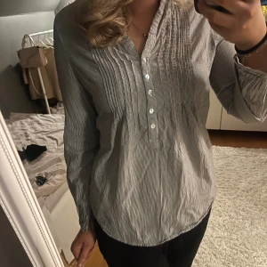 Randig blus - Suuper fin blus i gråvitt med tunna ränder. Blusen har v-ringning, dekorativa veck framtill och fyra blanka knappar.🍁🍂