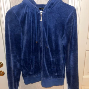 Blå velour hoodie från Juicy Couture - Mysig blå hoodie från Juicy Couture i mjuk velour med huva och dragkedja med glittrande J-hänge i guld. Klassisk modell med ribbade muddar och fickor framtill. Perfekt för en chill och trendig look. Säljs för ca 500kr på Depop, jag säljer för 150