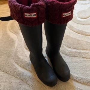 Hunter svarta stövlar med vinröda muddar - Hunter svarta höga stövlar i strl 40/41, men är mer 39/40. Baksidan har diskret rutigt mönster. Perfekta för regniga dagar och höstpromenader. Köpta för några år sedan i London! Kommer med ett par fina vinröda vinter strumpor 