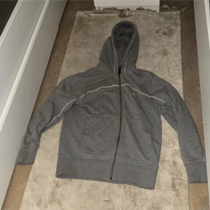 Grå Hugo boss hoodie med dragkedja och huva - Säljer en grå hoodie med hel dragkedja och huva. Tröjan har två stora fickor framtill och en tunn vit linje som detalj längs ärmar och bröst. Perfekt för chill dagar eller när du vill ha något bekvämt och enkelt. 