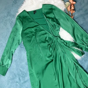 Grön omlottklänning med knyt - Snygg grön omlottklänning från Only  med långa ärmar och breda manschetter. Klänningen har ett elegant knyt i midjan och v-ringad hals. Materialet är mjukt och har en lätt glansig finish som ger en lyxig känsla. Perfekt för dig som vill sticka ut med färg.