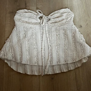 Beige tubtopp med spets från SHEIN - Supersöt beige tubtopp från SHEIN i storlek XS. Toppen har dekorativa spetsdetaljer och rynkade ränder som ger en romantisk vibe. Den är axelbandslös och har knyt framtill för extra stilpoäng. Perfekt för dig som gillar boho och vill ha något unikt i garderoben. Använd ca 3-4 gånger 🌟