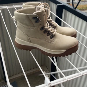 Beiga boots från Everest, storlek 38 - Snygga beiga boots från Everest med grov sula i brunt och snörning framtill. Skorna har en klassisk outdoor-look med robusta metalldetaljer och vadderad insida för extra komfort. Perfekta för höst och vinter när du vill ha både stil och funktion.