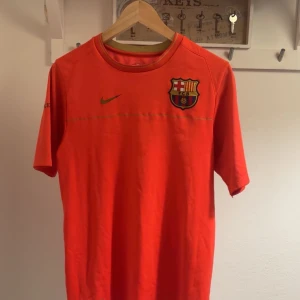 FC Barcelona Nike fotbollströja 2008/2009 - Säljer en FC Barcelona fotbollströja från Nike i starkt orange färg. Tröjan har klubbmärket på bröstet, Nike-logga och texten FC Barcelona på ärmen. Klassisk rund hals och korta ärmar, tillverkad i lätt och ventilerande material.