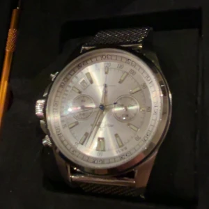 Jacques Lemans silverfärgad kronograf - Säljer en stilren kronograf från Jacques Lemans med silverfärgad urtavla och mesh-armband i metall. Medföljer även ett brunt läderarmband med krokodilpräglad yta och verktyg för byte. Klockan har flera subdials och datumvisning, levereras i original ask.