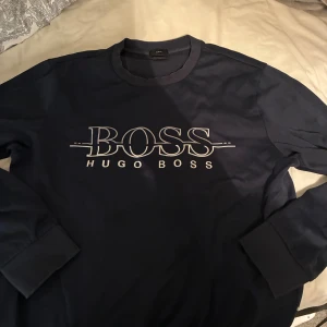 Marinblå Hugo Boss sweatshirt - Snygg marinblå sweatshirt från Hugo Boss med stort vitt logotryck på bröstet. Tröjan har rund hals, långa ärmar och ribbade muddar. Perfekt för dig som gillar stilrena och klassiska plagg med en modern touch.