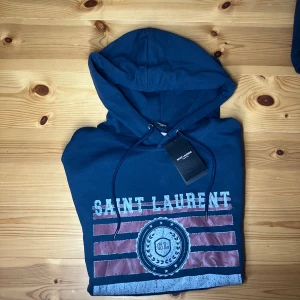 Marinblå Saint Laurent hoodie - Snygg marinblå hoodie från Saint Laurent med tryck framtill i vitt och rött, samt coola detaljer som snörning i huvan och känguruficka. Tröjan har lång ärm och är tillverkad i mjuk bomull, perfekt för en avslappnad och trendig look.