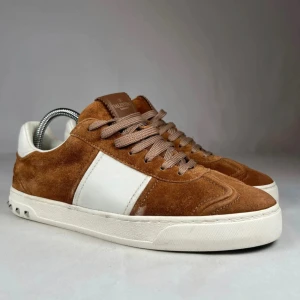 Valentino Garavani bruna sneakers mocka - Snygga sneakers från Valentino Garavani i brun mocka med vita skinnpaneler och vita detaljer. Skorna har platt sula med nitar baktill och bruna snören. Klassisk rund tå och coolt mönstrad yttersula. Perfekta för dig som gillar exklusiva och stilrena sneakers.