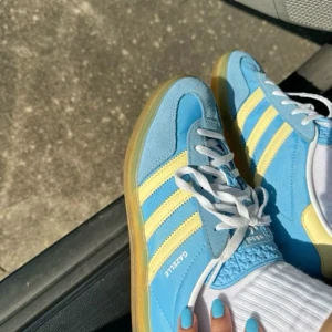 Adidas Gazelle blå sneakers strl 39 - Fräscha Adidas Gazelle sneakers i blått med ljusgula ränder och detaljer. Skon har en klassisk låg siluett, vit snörning och en genomskinlig gummisula. Ovandelen är i mocka och syntet, perfekt för dig som gillar retrostil och färg. Nyskick