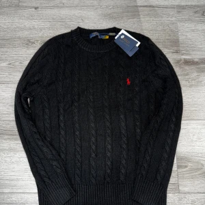 Svart kabelstickad tröja Polo Ralph Lauren - Svart kabelstickad tröja från Polo Ralph Lauren med klassisk rund hals och röd broderad logga på bröstet. Tröjan har långa ärmar och ribbade muddar vid ärmslut och nederkant. Perfekt för dig som gillar stilren och tidlös design. Ete kvalité 