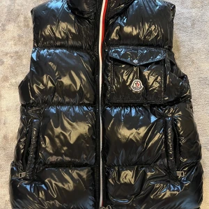 Svart dunväst från Moncler - Snygg svart dunväst från Moncler med glansig finish och quiltad design. Västen har en bröstficka med Moncler-logga, två sidofickor med dragkedja och en klassisk ståkrage. Perfekt för lager-på-lager och streetstyle. Fodrad med vitt gåsdun för extra värme.