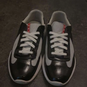 Prada sneakers svart och vit mesh - Säljer ett par snygga Prada sneakers med svart lackad skinnpanel och vit mesh. Skorna har vita snören, rund tå och en chunky vit sula. Röd Prada-logga syns på plösen och hälen. Perfekt för dig som gillar exklusiva och sportiga vibes.Pris går att diskutera via en snabb och smidig affär.