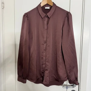 Brun skjorta med knappar från Vila - Snygg lila-/burgundy-färgad skjorta från Vila i mjuk, glansig polyester. Klassisk krage, dolda knappar framtill och puffade axlar ger en cool touch. Långärmad med knapp vid manschetten. Perfekt för dig som gillar stilrena plagg med lite extra detaljer.