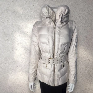 Beige pufferjacka med bälte - Snygg beige pufferjacka med  markerad midja tack vare det breda bältet med metallspänne. Jackan har quiltad design, dragkedja framtill och två fickor med zip. PS: Liten reva på insidan, påverkar inte användningen # Zara #vintjackor