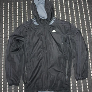 Svart vindjacka från Adidas - Svart vindjacka från Adidas med huva och hel dragkedja framtill. Jackan har diskreta grå detaljer på axlarna och baksidan, samt två sidofickor med dragkedja. Perfekt för blåsiga dagar och sportiga outfits. Adidas-logga på bröstet och Ultra Proof-tryck baktill.