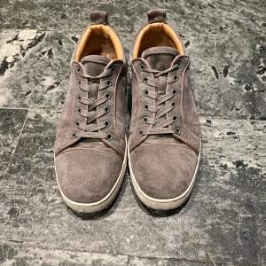 Louboutin skor - Säljer ett par gråa sneakers från Christian Louboutin i mocka med klassisk röd sula och snörning. Skorna har rund tå, vita kanter och beige insida. Perfekta för dig som vill ha en stilren men ändå lyxig look.