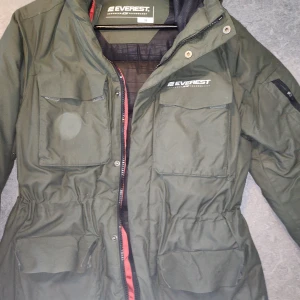 Grön parkajacka från Everest, strl 38 - Mörkgrön parkajacka från Everest med flera stora fickor framtill, dragsko i midjan och svart innerfoder. Jackan har dragkedja och tryckknappar samt diskreta röda detaljer längs insidan. Perfekt för kyliga dagar.
