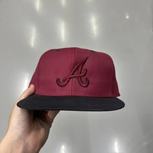 Vinröd Atlanta Braves keps - Snygg vinröd snapback-keps med svart broderad A-logga framtill och svart skärm. Undersidan av skärmen är grön för extra kontrast. Klassisk rak modell som passar streetwear-stilen. Perfekt för dig som vill ha en sportig look.