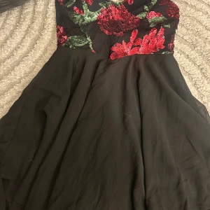 Svart kort klänning med paljettblommor - Supersnygg svart kort klänning med axelbandslös överdel täckt av röda och gröna paljettblommor. Kjolen är i flera lager tunt tyg som ger ett flowy intryck. Perfekt för dig som vill sticka ut med glittriga detaljer och blommigt mönster.