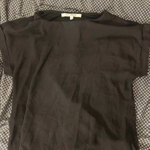 Svart blus från KappAhl, storlek S - En stilren svart blus från KappAhl i storlek S. Blusen har en enkel, rak passform med korta ärmar och rund halsringning. Materialet känns mjukt och har en lätt glans, vilket ger en snygg look. Perfekt för dig som gillar minimalistisk stil.