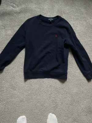 Mörkblå sweatshirt från Polo Ralph Lauren - Snygg mörkblå sweatshirt från Polo Ralph Lauren i storlek L. Tröjan har ett klassiskt crewneck-utseende och en liten röd broderad logga på bröstet. Tillverkad i mjuk bomull med långa ärmar och ribbade muddar vid hals, ärmslut och nederkant.