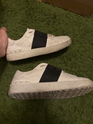 Valentino Garavani vita sneakers - Säljer ett par snygga vita Valentino Garavani sneakers med bred svart rem över sidan. Skorna har platt sula med mönstrad undersida och coola nitar längs kanten. Tillverkade i skinn med snörning och rund tå. Perfekt för dig som gillar stilrena designer.