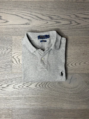 Grå långärmad polotopp från Polo Ralph Lauren - Snygg grå långärmad polotopp från Polo Ralph Lauren i slim fit. Klassisk krage, två knappar framtill och ikonisk broderad logga på bröstet. Mjuk bomullskvalitet som känns skön mot huden. Perfekt för dig som gillar stilrena och tidlösa plagg.
