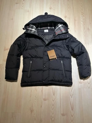 Svart pufferjacka Burberry - Säljer en svart pufferjacka från Burberry med klassiskt rutigt foder i huvan. Jackan har dragkedja, knappar och två fickor framtill. Perfekt för kalla dagar och har en snygg, modern look med diskret Burberry-logga på ärmen.