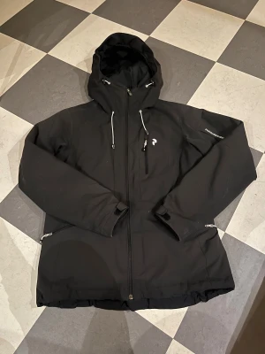 Peak performance jacka - Svart jacka från Peak Performance med huva och snörning, dragkedja framtill och praktiska fickor med dragkedja. Jackan har en stilren look och är perfekt för kyliga dagar. Snygg logga på bröstet och diskret branding på ärmen. Jackan funkar perfekt för att åka skidor eller bara ha som vardagsjacka.
