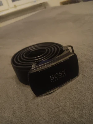 Svart Hugo Boss skärp i skinn - Snyggt svart skärp från Hugo Boss med stilren metallspänne och präglad logga. Skärpet är tillverkat i äkta skinn med diskreta sömmar längs kanterna och har en modern, klassisk look. Perfekt accessoar för att lyfta din outfit.