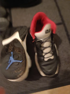 Nike Jordan sneakers svart/vit/röd - Nike Jordan sneakers med svart och vit ovandel, röda detaljer på insidan och blå Jumpman-logga på sidan och sulan. Skorna har snörning och en rund tå, tillverkade i skinn och syntet. Snygg kontrast mellan färgerna och klassisk Jordan-design.