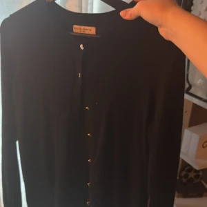 Svart kofta från Holly & Whyte - Svart, klassisk kofta från Holly & Whyte by Lindex i storlek S. Koftan har rund hals, långa ärmar och snygga knappar framtill. Materialet känns mjukt och lätt, perfekt för lager-på-lager. Enkel och stilren design som funkar till allt.