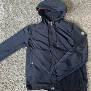 Mörkblå vindjacka från Moncler - Snygg mörkblå vindjacka från Moncler med huva och dragsko. Jackan har dragkedja framtill, två fickor med tryckknapp och Moncler-logga på ärmen. Perfekt för blåsiga dagar och enkel att matcha med det mesta. Snabbaffär först till kvarn, för mer bilder PM!