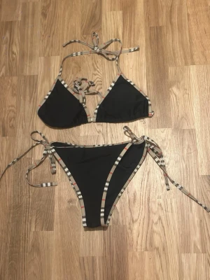 Svart bikini med rutiga band, strl L - Padsen i toppen kan tas bort om det önskas. Toppen har trekantsform och knyt i nacken och ryggen, medan trosan har smala band på sidorna. Materialet är 80% polyester och 20% elastan.