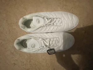 Nike Air Max Plus vita sneakers - Nike Air Max Plus sneakers i helvit färg med snygga vågiga detaljer och synliga luftkuddar i sulan. Skorna har snörning, rund tå och en platt sula med svarta och röda inslag undertill. Materialet är syntet och mesh för en fräsch och sportig look.