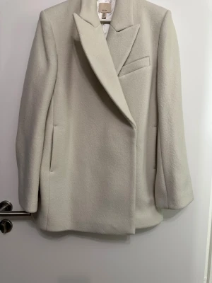 Beige ullkappa från H&M XS - Stilren beige kappa/kavaj från H&M i ullblandning i stl xs. Klassisk design med bred krage, dolda fickor och rak passform. Kappa/kavajen är i tjockare material. Helt ny aldrig använd. 