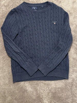 Mörkblå kabelstickad tröja GANT - Snygg mörkblå kabelstickad tröja från GANT i premium bomull. Klassisk rund hals och ribbade muddar vid ärmslut och nederkant. GANT-logga broderad på bröstet. Perfekt för dig som gillar stilrena och tidlösa plagg. Tröjan är i bra skick inga fläckar den är väl använd. Pm för mer frågor😊