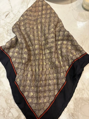 Gucci sidenscarf - Snygg fyrkantig scarf från Gucci i brunt och beige med klassiskt GG-mönster och gulddetaljer. Svart kant med röd linje ger en lyxig touch. Perfekt att styla runt halsen eller som accessoar. Materialet är mjukt siden.