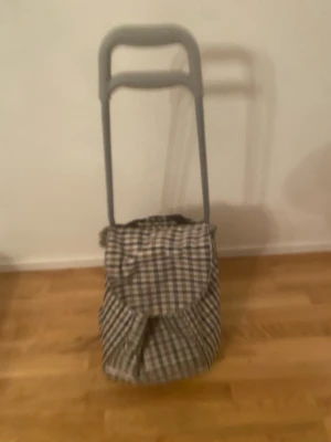 Rutig shoppingväska med hjul Dramaten  - Praktisk shoppingväska med två hjul och grått handtag. Väskan har ett rutigt mönster i beige, brun och svart och är tillverkad i ett slitstarkt tygmaterial. Perfekt för att rulla hem matvaror eller packa inför en dag på stan.