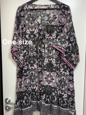 Blommig boho-standklänning med volang - Superfin boho-inspirerad strandklänning med blommigt mönster i svart, rosa och vitt. StrandKlänningen har v-ringning med knyt, trekvartsärmar med rosa spetsdetalj och volang nertill. Perfekt för dig som gillar avslappnad och trendig stil.🌸Använd två gånger. Storlek one size.