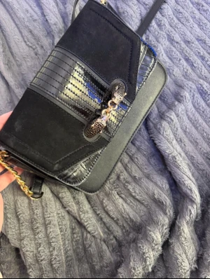 Svart axelväska från topchop  - Snygg svart axelväska med mix av mocka och skinn, detaljerad med glansiga partier och en cool kedja framtill. Väskan har en stilren rektangulär form och axelrem i kombination med guldfärgad kedja. Perfekt för dig som gillar att sticka ut med accessoarer.