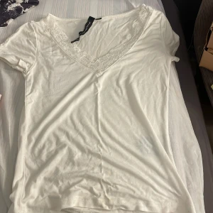 Vit v-ringad topp med spets - Säljer en vit v-ringad topp med fina spetsdetaljer runt halsringningen. Toppen har korta ärmar och är gjord i mjuk bomull, perfekt för en chill och stilren look. Spetsen ger en extra touch av elegans och feminin vibe.