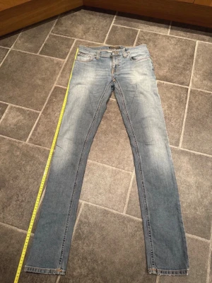 Nudie Jeans ljusblå slim fit jeans W27 L34 - Säljer ett par ljusblå skinny jeans från Nudie Jeans med klassisk femficksdesign och orangea kontrastsömmar. Jeansen har normal passform i midjan och är tillverkade i mjukt jeanstyg. Perfekt för dig som gillar en smal siluett och stilren look. Längd 106 cm bredd 38 cm