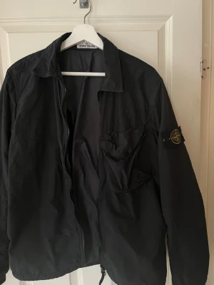 Svart overshirt från Stone Island - Svart overshirt från Stone Island med klassisk krage, dragkedja framtill och ikonisk patch på vänster ärm. Jackan har en stor ficka med knappstängning på bröstet och är tillverkad i lätt polyester. Perfekt för dig som vill ha en clean och stilren look.