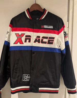 Race bomberjacka från United Racing Society - Snygg bomberjacka med racing-inspirerad design från United Racing Society. Jackan har svarta, vita, röda och blå paneler, tryck med 'X RACE', '1972' och schackrutiga flaggor. Ribbad krage och muddar med röd och vit detalj. Materialet är blankt och känns sportigt.