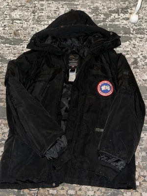 Svart dunjacka från Canada Goose - Svart dunjacka från Canada Goose med huva och klassisk patch på bröstet. Jackan har flera fickor, ribbade muddar och dragkedja framtill. Materialet är slitstark polyester och dunfyllning, perfekt för kalla vinterdagar. Snygg broderad logga på ryggen.