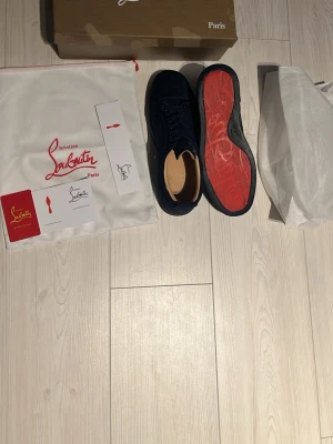 Mörkblå sneakers från Christian Louboutin - Säljer ett par snygga mörkblå sneakers från Christian Louboutin med ikonisk röd sula. Skorna har rund tå, snörning och är tillverkade i mocka för en lyxig känsla. Perfekta för dig som vill sticka ut med exklusiv stil. 1000 kr så är dom dina!